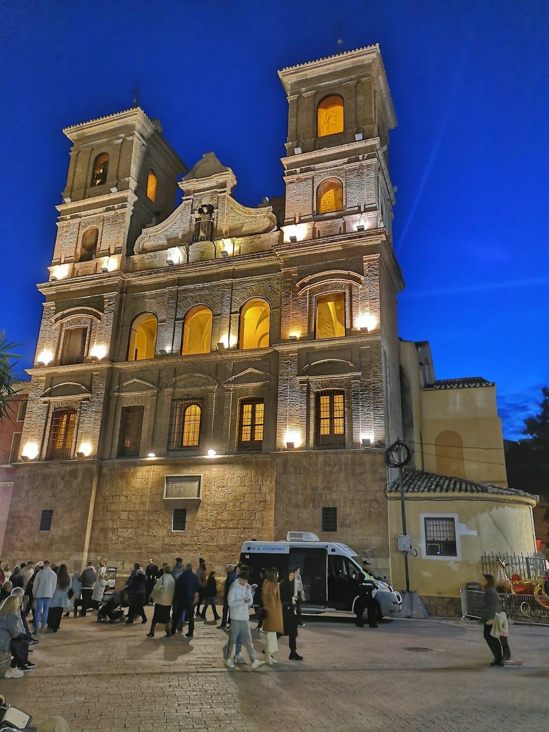 Persone riunite in una piazza acciottolata davanti a una chiesa illuminata.